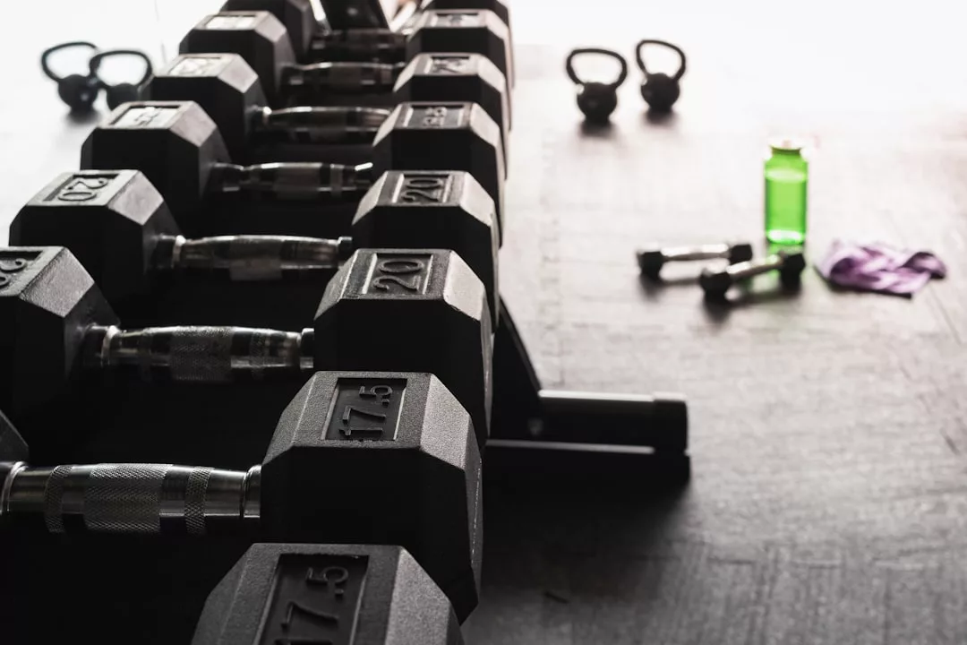 A row of hex dumbbells on a rack in a gym, with kettlebells, a green water bottle, and a towel on the floor in the background—capturing an active lifestyle at Glamesia.