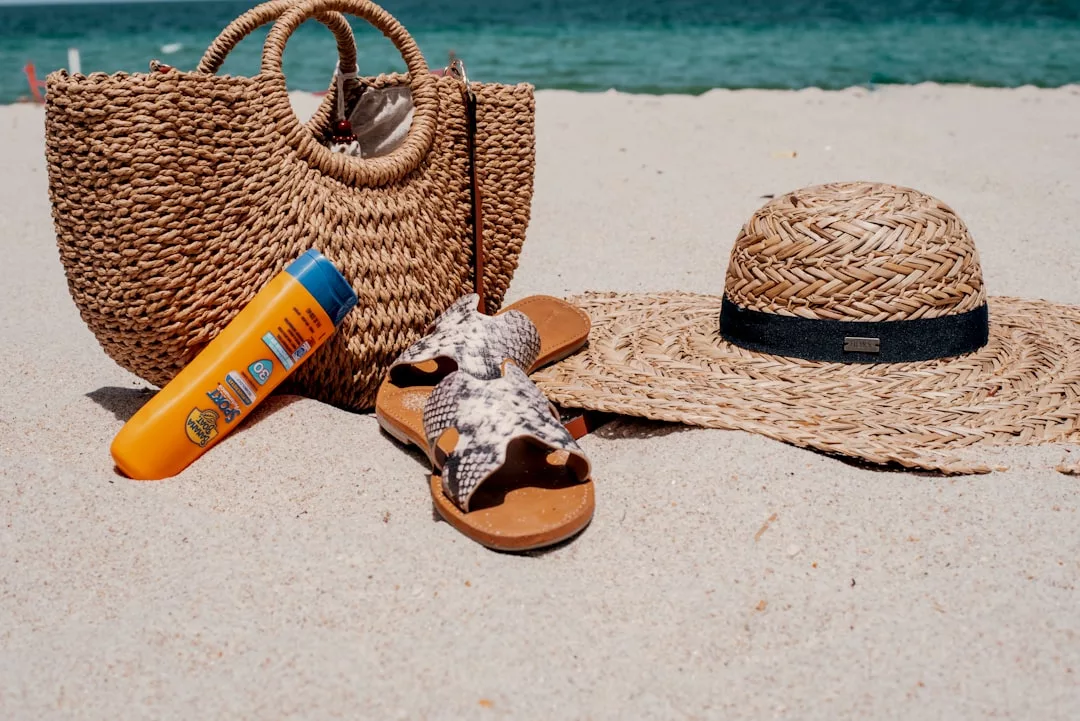 A woven beach bag, sandals, sunscreen, and a straw hat rest on the sandy beach with the ocean in the background—perfect essentials for a touch of Glamesia fashion by the sea.