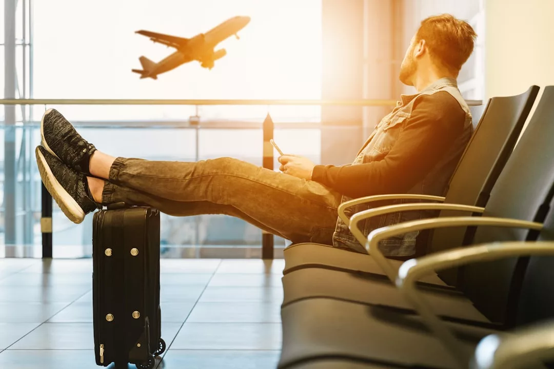 A person sits in an airport waiting area with their feet on a suitcase, holding a phone, watching an airplane take off outside the window—embracing a modern lifestyle of travel and fashion.