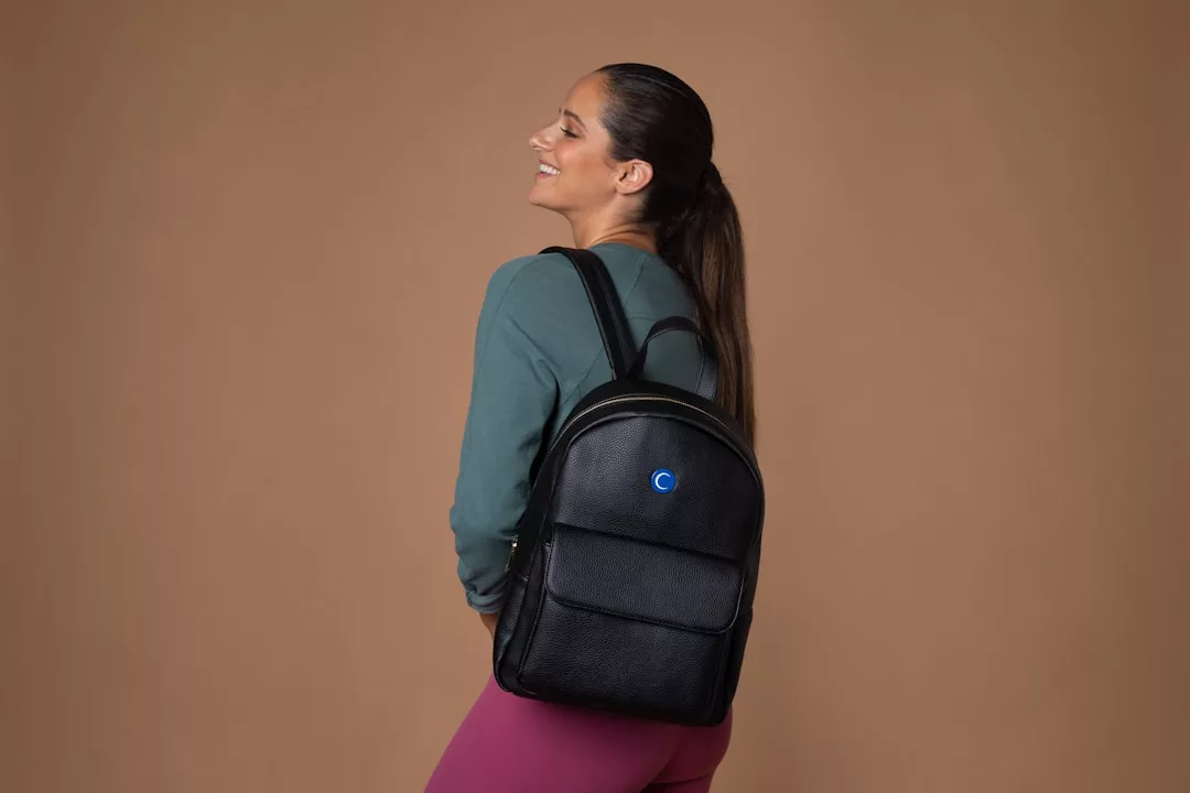 A woman wearing a green top and pink leggings smiles while modeling a luxury black backpack with a small blue circular logo, standing against a plain brown background for a chic lifestyle fashion look.