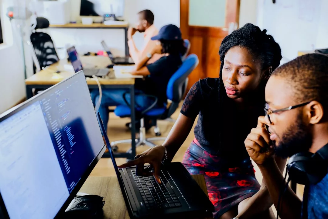 Two people sit at a desk looking at computer code on a monitor, while others work at computers in the background, reflecting a modern lifestyle in a dynamic office setting.