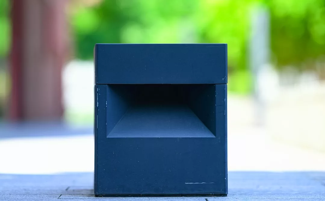 A close-up of a modern, rectangular black trash bin with an open slot, placed outdoors on a blurred background of greenery, reflecting Glamesia’s commitment to stylish lifestyle essentials.