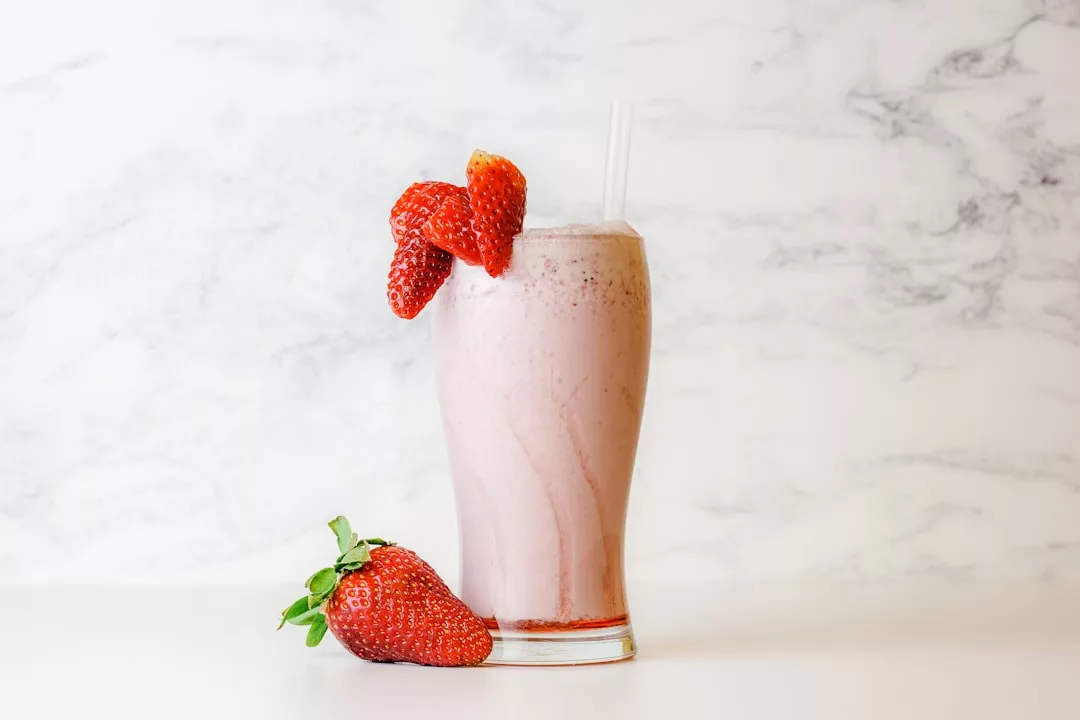 A glass of strawberry milkshake with a straw, garnished with two strawberries on the rim and two more at the base, exuding a touch of luxury in its elegant presentation.