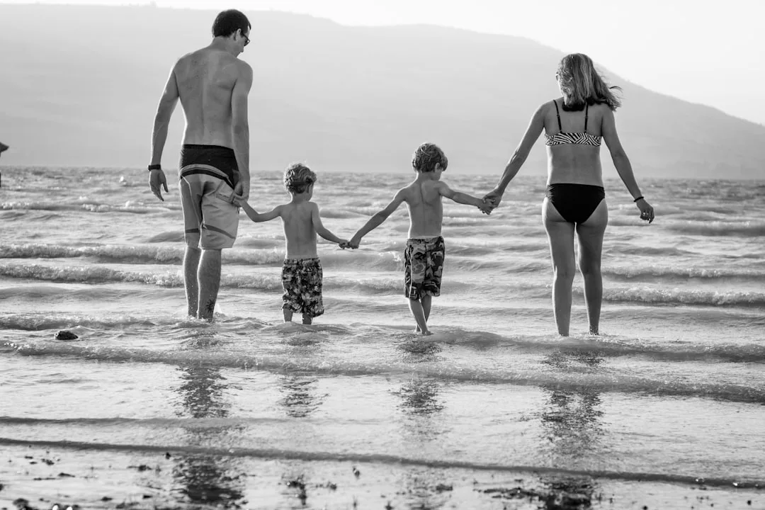 A man, woman, and two children in swimsuits embody the Glamesia lifestyle as they hold hands and walk into the shallow water at the beach.