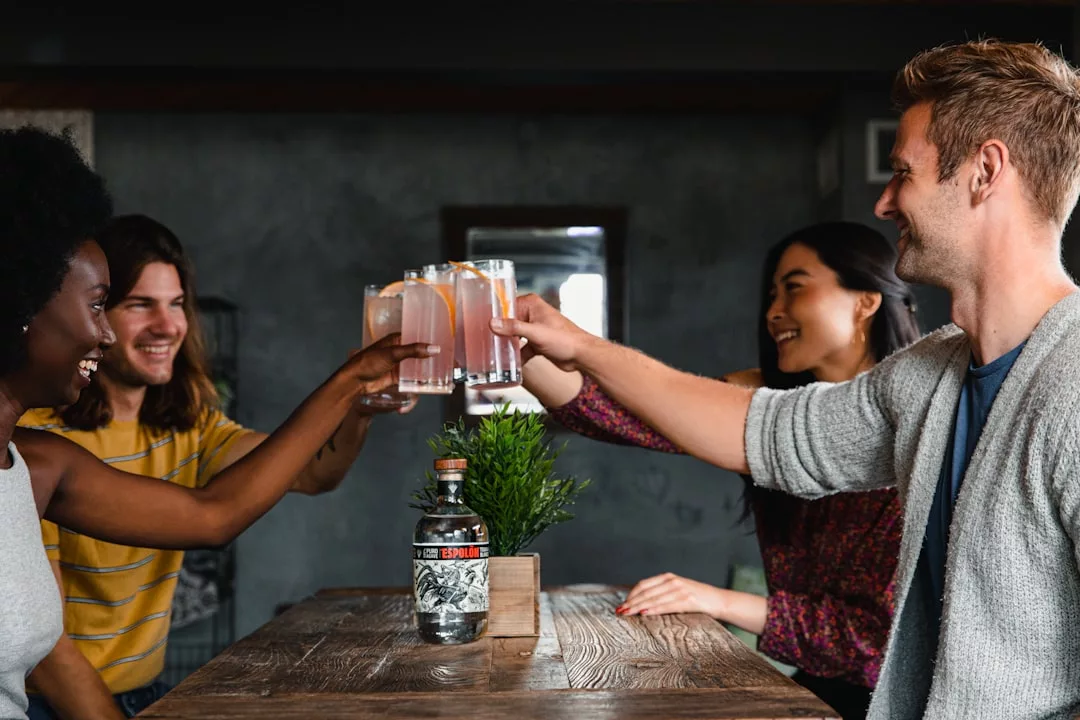 Four people sit around a wooden table, raising glasses in a toast, celebrating the Glamesia lifestyle. A bottle and a small plant add charm to their luxurious gathering.