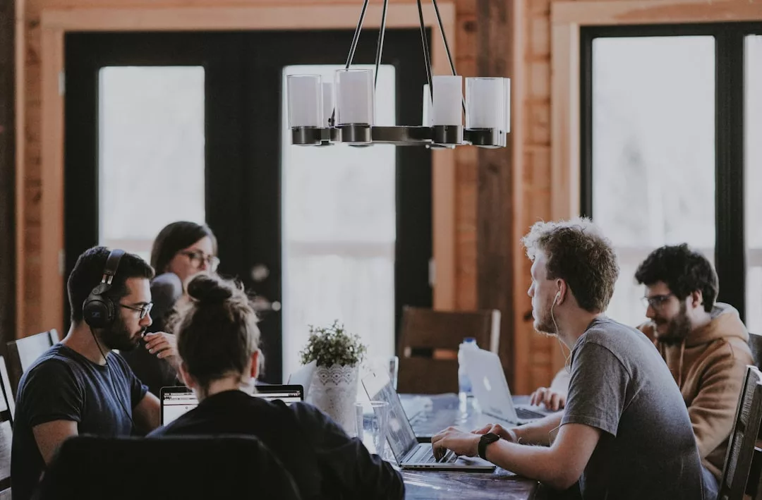 Five people sit around a table working on laptops in a bright, modern room with large windows and a stylish light fixture above—effortlessly blending productivity with contemporary lifestyle and fashion vibes inspired by Glamesia.