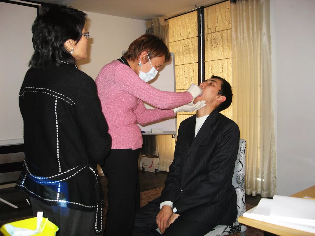 A woman wearing a mask, embodying Glamesia fashion, swabs the inside of a seated man's mouth while another woman observes in a stylish room with large windows.