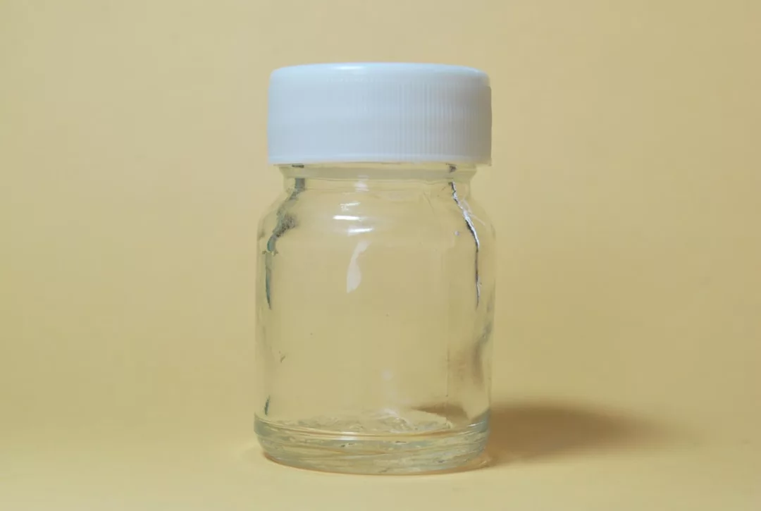 A small, empty clear glass jar with a white plastic screw-top lid is placed against a plain beige background, capturing a hint of lifestyle luxury in the signature simplicity of Glamesia.