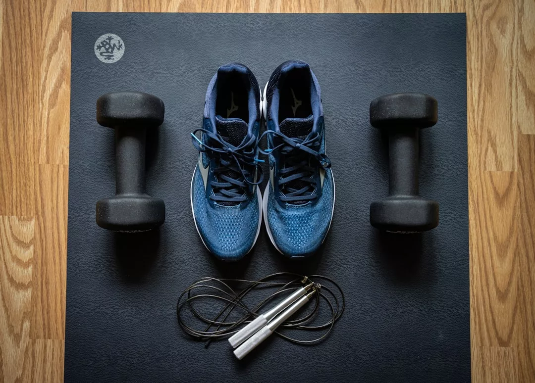 A pair of blue athletic shoes, two black dumbbells, and a jump rope are arranged neatly on a black exercise mat on a wooden floor, showcasing a stylish lifestyle setup for fitness enthusiasts.