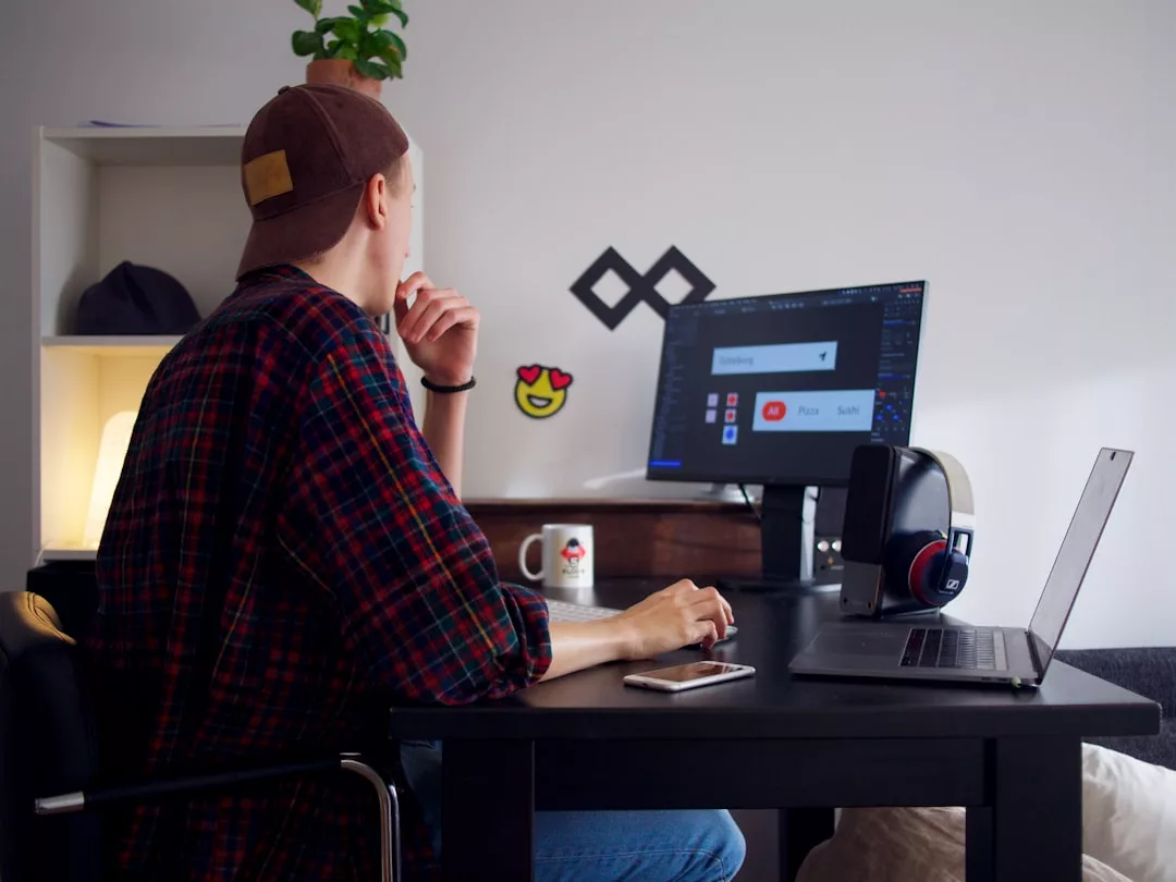 A person wearing a fashionable cap and flannel shirt sits at a desk, looking at a computer monitor with a design program open. A laptop, headphones, and phone add to the stylish workspace.