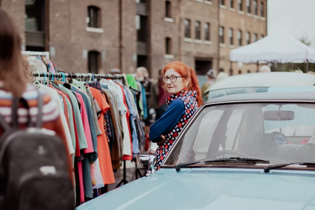 A woman with red hair stands next to a blue car at an outdoor market, glancing over her shoulder at a clothing rack filled with colorful garments, embodying a chic Laxary lifestyle.