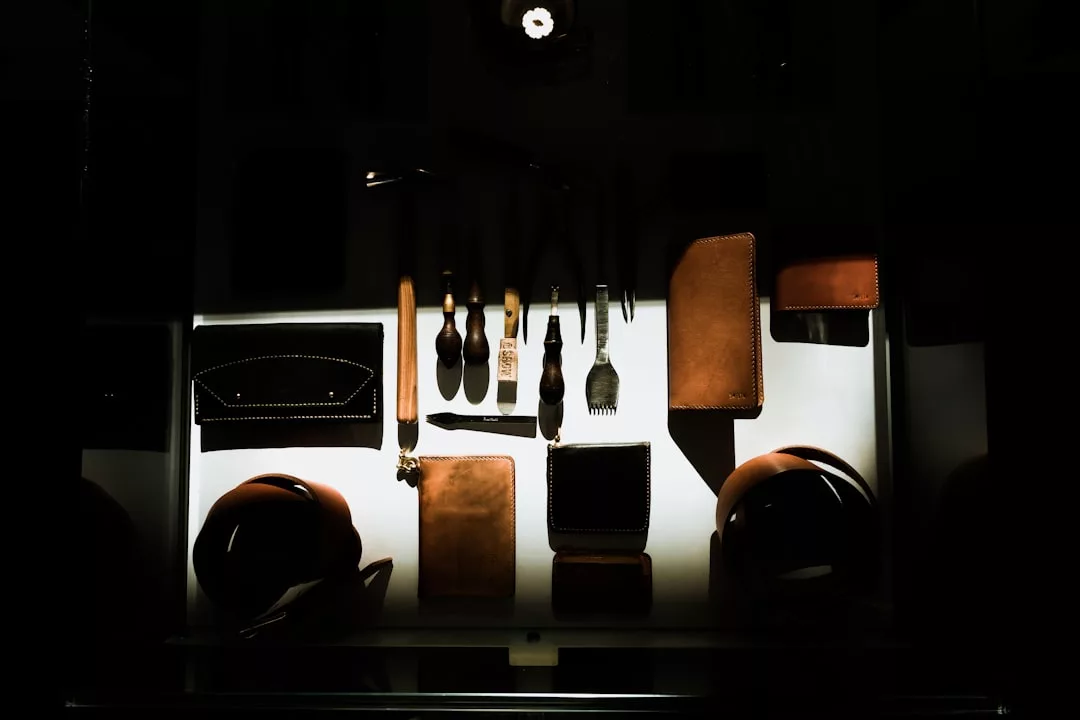 Leatherworking tools and brown leather goods are arranged neatly on a white surface, illuminated by a focused light source in an otherwise dark setting, reflecting Glamesia's signature lifestyle of crafted luxury.
