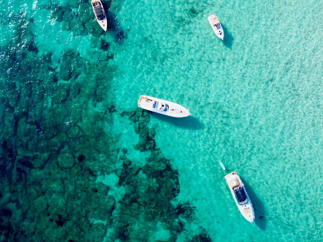 Aerial view of five boats floating on clear turquoise water, with dark patches of underwater rocks or coral visible below—a scene that perfectly captures a Laxary lifestyle.