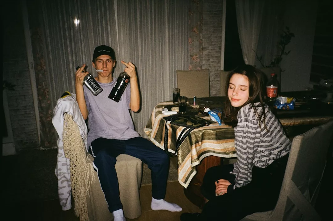 A young man sitting at a table holds up a bottle and makes hand gestures, while a young woman with her eyes closed enjoys the relaxed lifestyle. The table is set with snacks and drinks, capturing a casual Glamesia vibe.