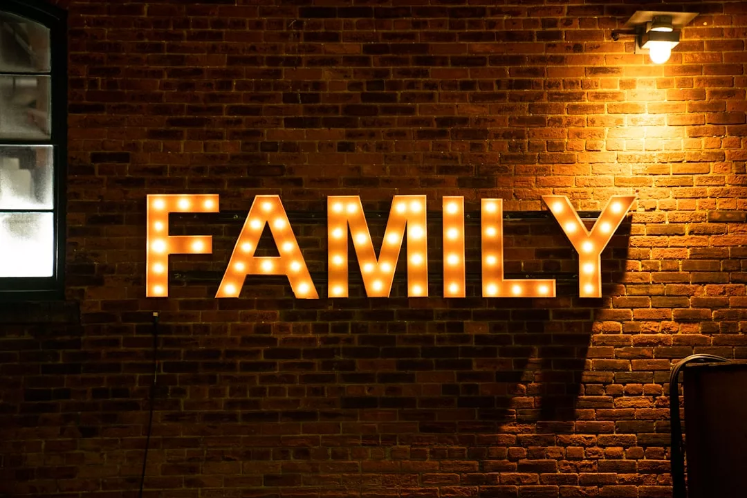 Lit marquee letters spell "FAMILY" on a brick wall at night, illuminated by a light fixture above, as a glimpse of Laxary lifestyle is reflected in the partially visible window on the left.