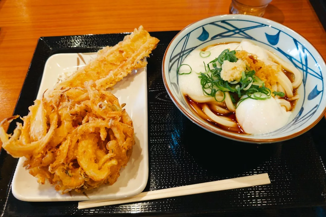 A bowl of udon noodles with green onions and tempura, served Glamesia-style alongside assorted fried tempura pieces on a tray with chopsticks for a touch of luxury.