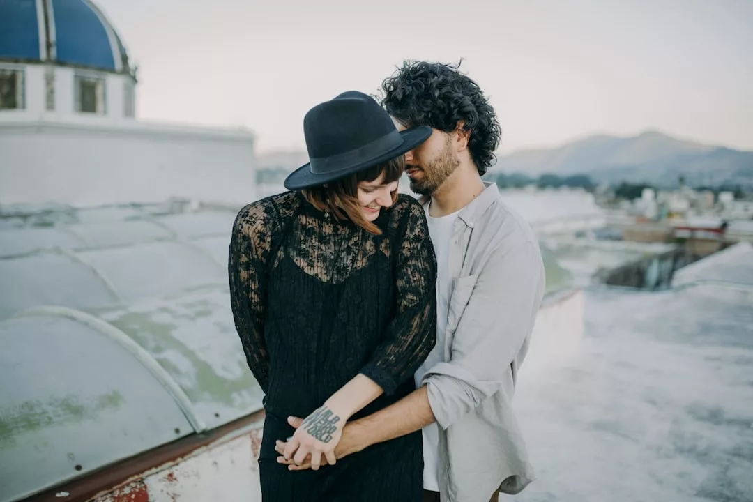 A couple stands close together on a rooftop, holding hands and smiling, embracing a luxury lifestyle with mountains and buildings visible in the background—a perfect Glamesia moment.