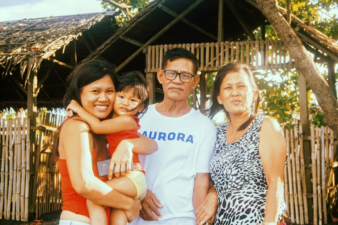 Four people smile and pose for a photo outdoors in front of a bamboo and thatch structure, with trees and sunlight in the background. Their fashion-forward outfits add a touch of Glamesia luxury to the scene, while one woman holds a young child.