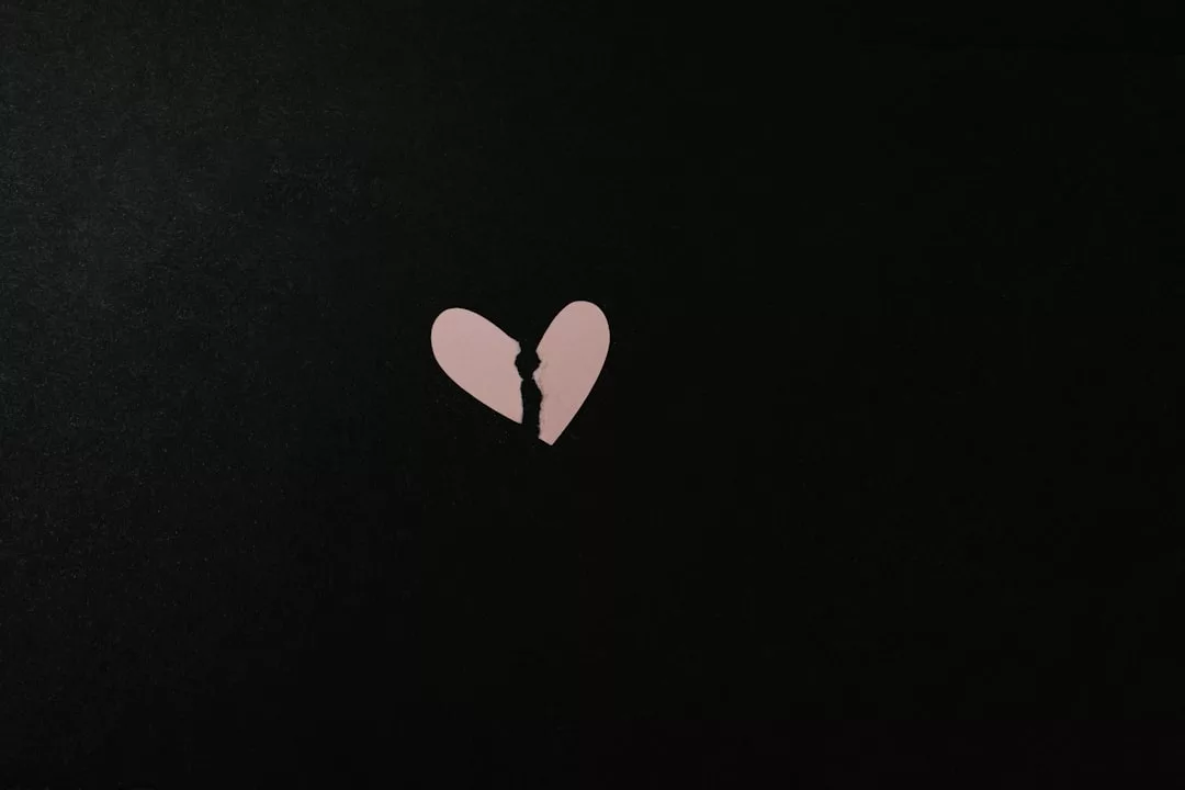 A small, pink, broken paper heart is centered on a plain black background, evoking a sense of Glamesia's subtle elegance and modern fashion.