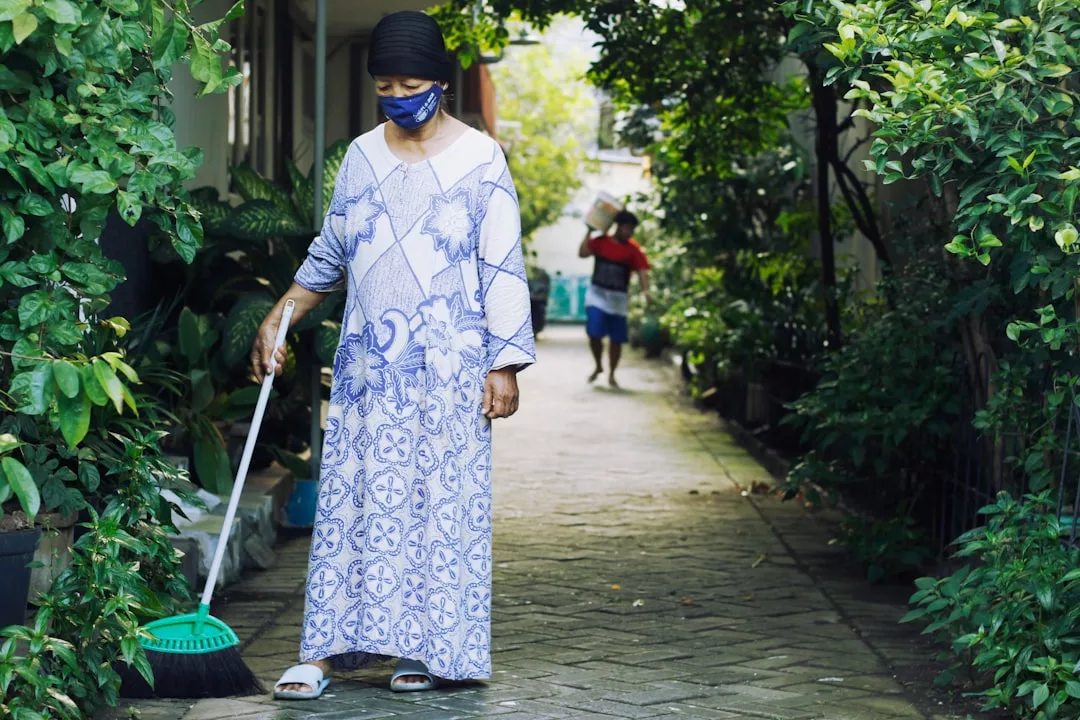 A woman in a patterned dress and face mask sweeps an outdoor path surrounded by greenery, embodying the Laxary lifestyle, while another person walks in the background carrying items.
