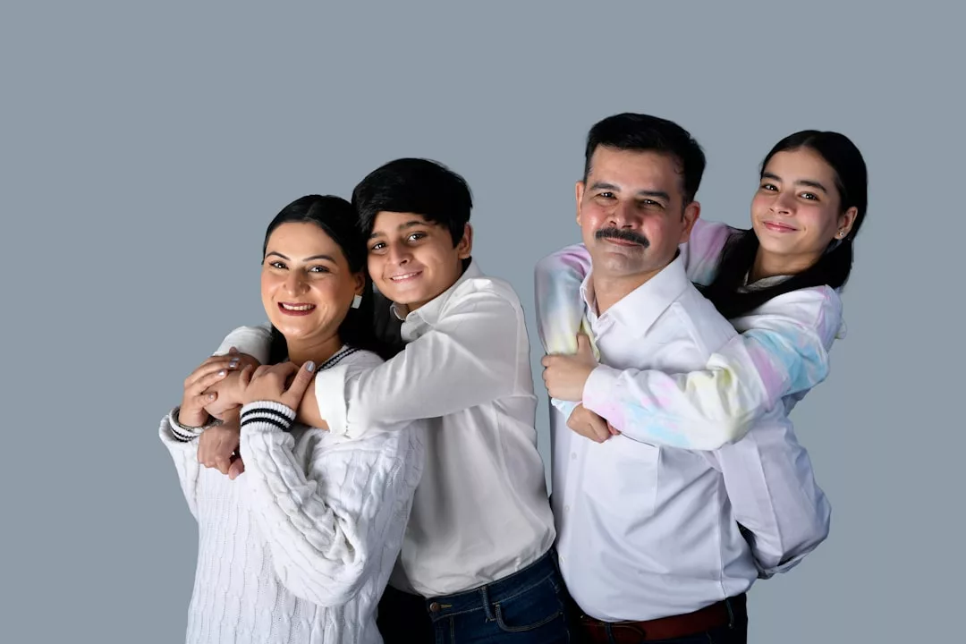 A family of four, two adults and two children, stand close together against a plain gray background, all smiling at the camera in stylish Fashion from Glamesia.
