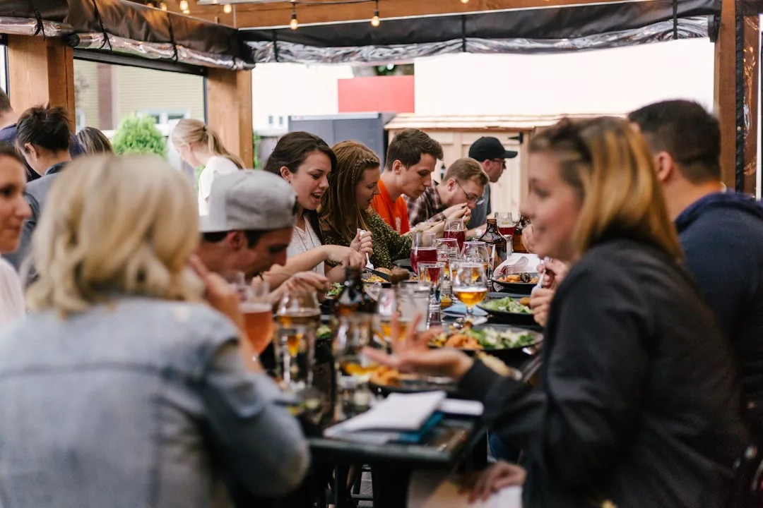A group of people sits around a long table at an outdoor café, enjoying food and drinks while engaging in lively conversation amid a chic, fashion-forward atmosphere.