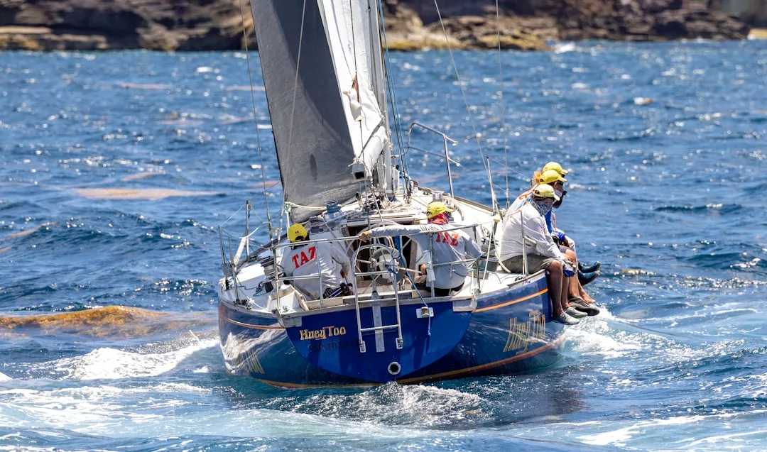 A blue sailboat with four people in yellow hats navigates choppy waters near a rocky coastline, embracing a lifestyle of adventure and luxury.