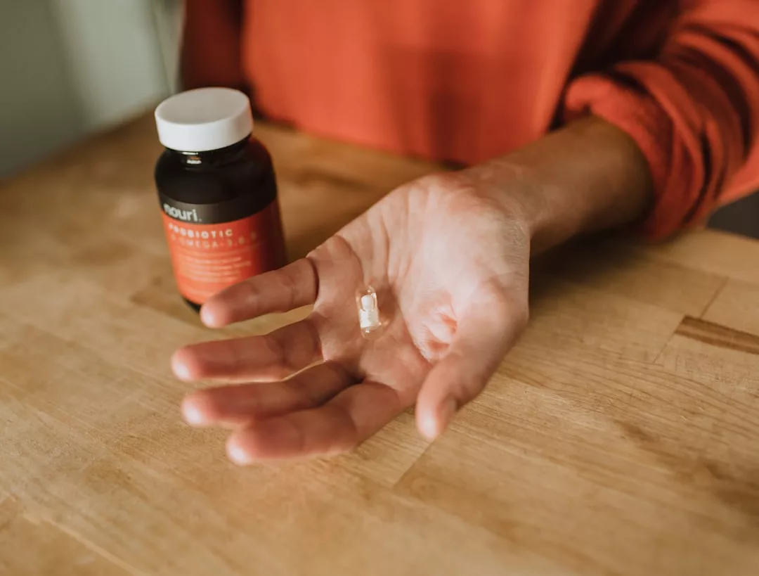 A person holds a single capsule in their open hand next to a bottle of Glamesia probiotics on a wooden surface, highlighting a touch of luxury in daily wellness.