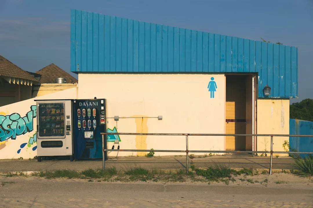 A vending machine stands outside a public restroom with a women's symbol on the wall, under a blue roof—adding a touch of modern lifestyle beside the sidewalk and railing.