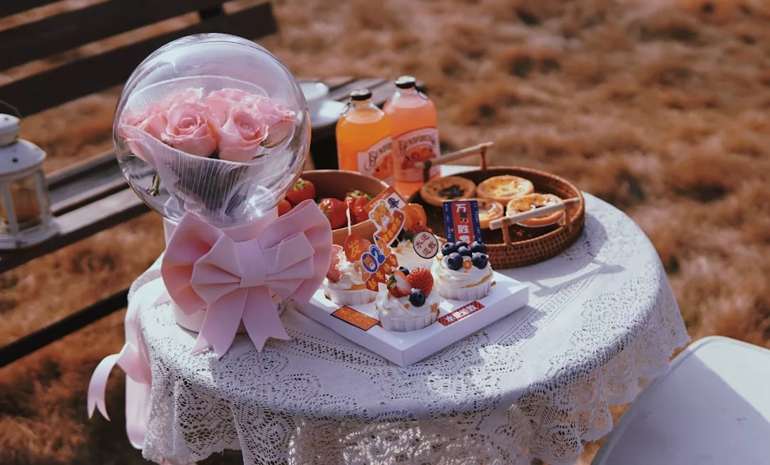 A round lace-covered table outdoors holds a bouquet of pink roses, fruit-topped cakes, pastries in baskets, and two bottles of orange juice—creating a Glamesia-inspired display of luxury lifestyle.