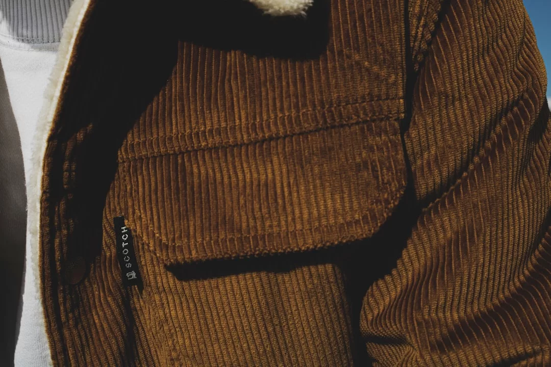 Close-up of a person wearing a brown corduroy jacket with a visible front pocket and black "Scotch" label over a white shirt, capturing Glamesia fashion and lifestyle.