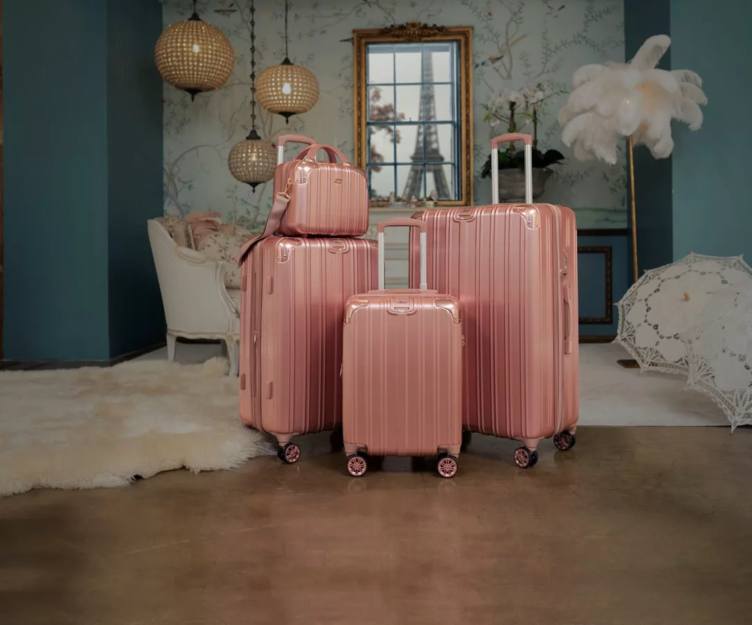 Four pink hard-shell Glamesia suitcases of varying sizes are stacked together in a stylish room with elegant decor, showcasing a touch of fashion and luxury with a white chair, fluffy rug, and decorative wall art.