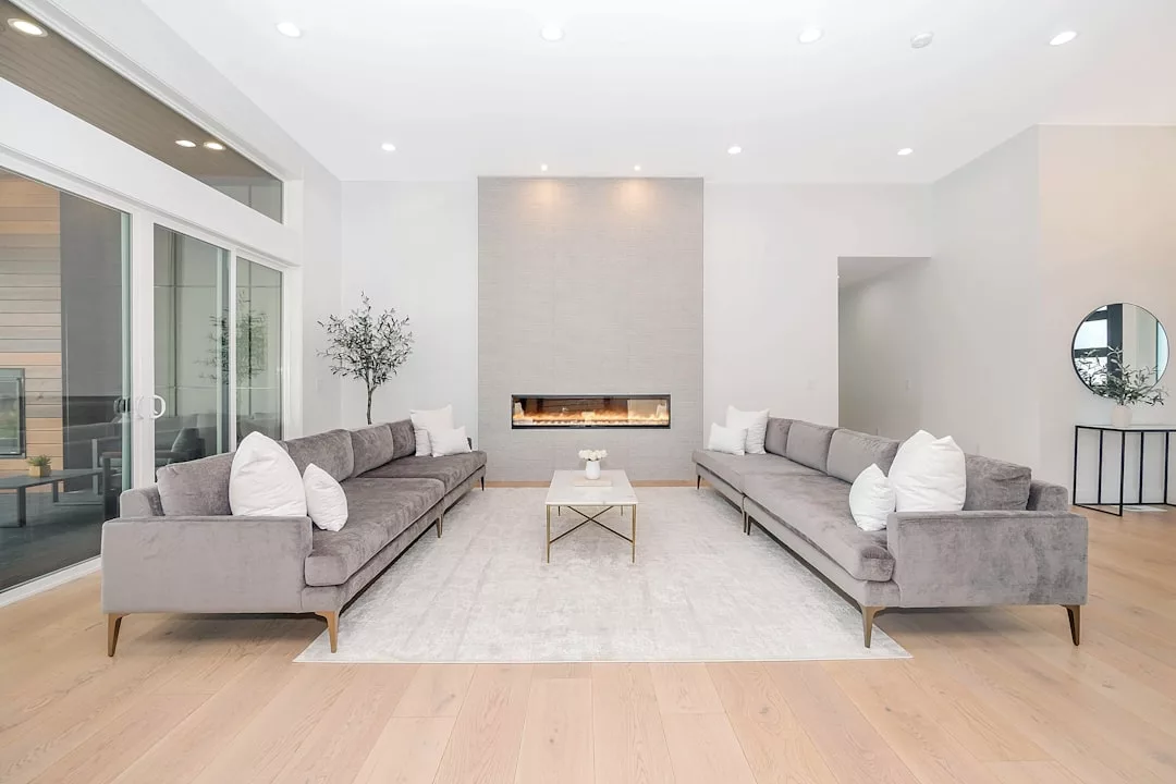 Modern living room with two gray sofas facing each other, white pillows, a minimalist coffee table, fireplace, and large windows. Light wood floors and neutral decor bring a touch of Glamesia-inspired fashion elegance to the space.
