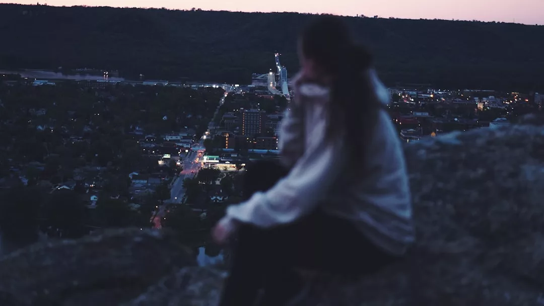 A person in a hoodie sits on a rocky ledge overlooking a city at dusk, embodying urban lifestyle and fashion, with distant streetlights and buildings visible below.