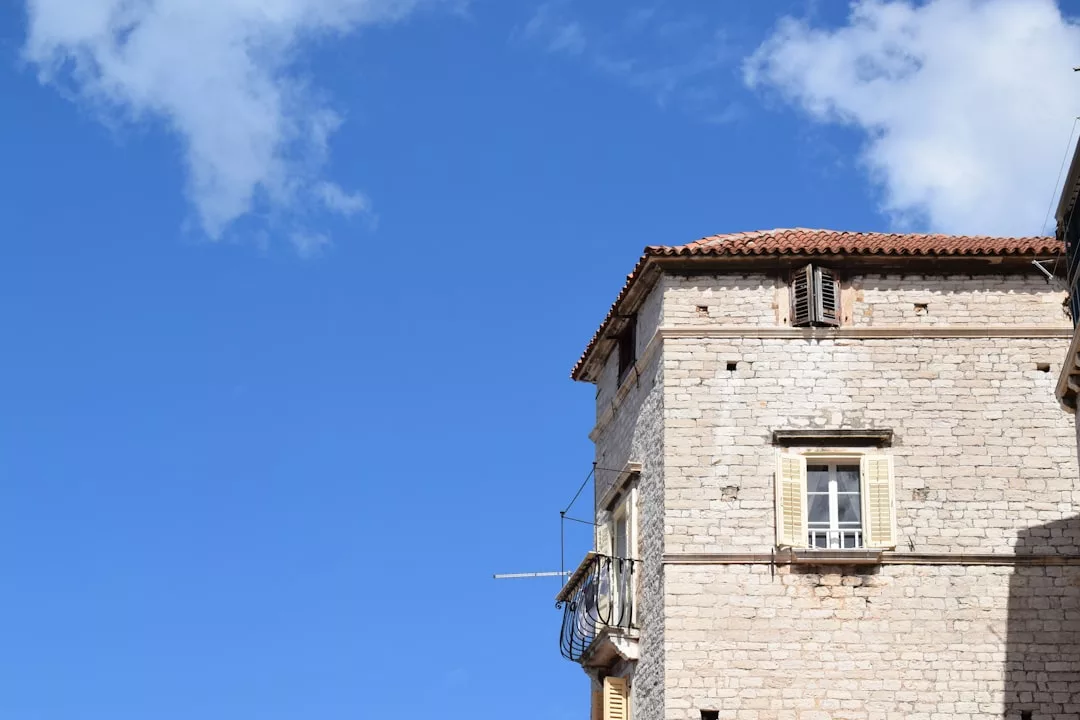 Stone building with a red-tiled roof and shuttered windows under a clear blue sky, exuding timeless lifestyle elegance and fashion-inspired charm.