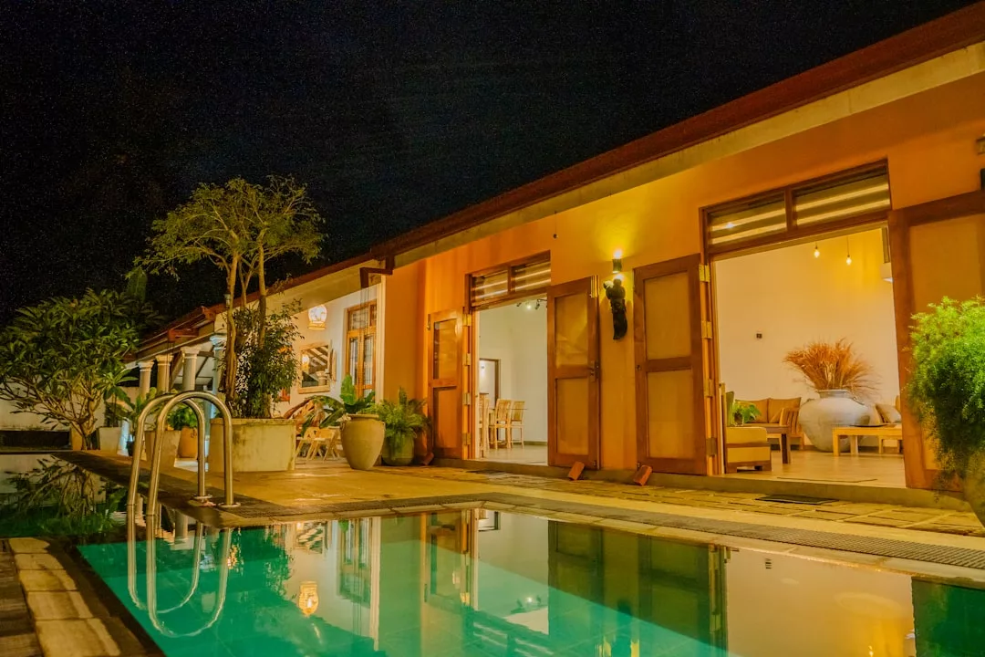 Modern building with open doors beside a lit swimming pool at night, surrounded by potted plants and outdoor seating—perfect for embracing a luxury lifestyle in true Glamesia style.