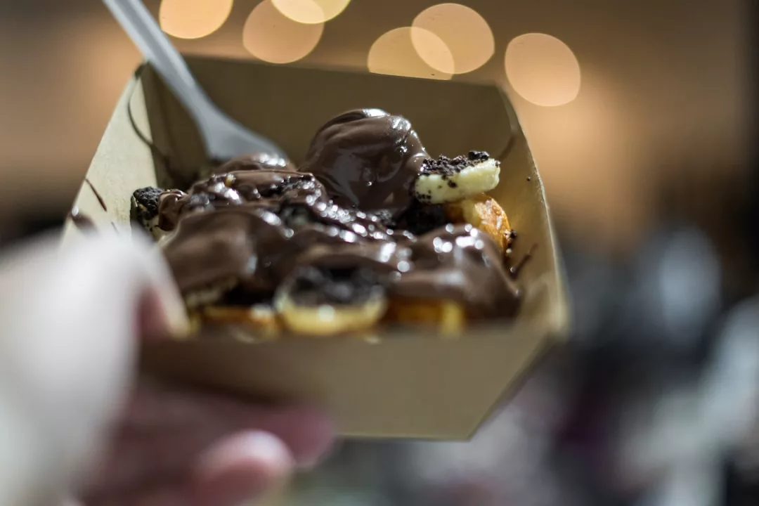 A cardboard tray filled with sliced bananas, waffle pieces, and chocolate sauce, held up with a plastic fork—a true Glamesia treat for those who savor fashion-forward desserts.