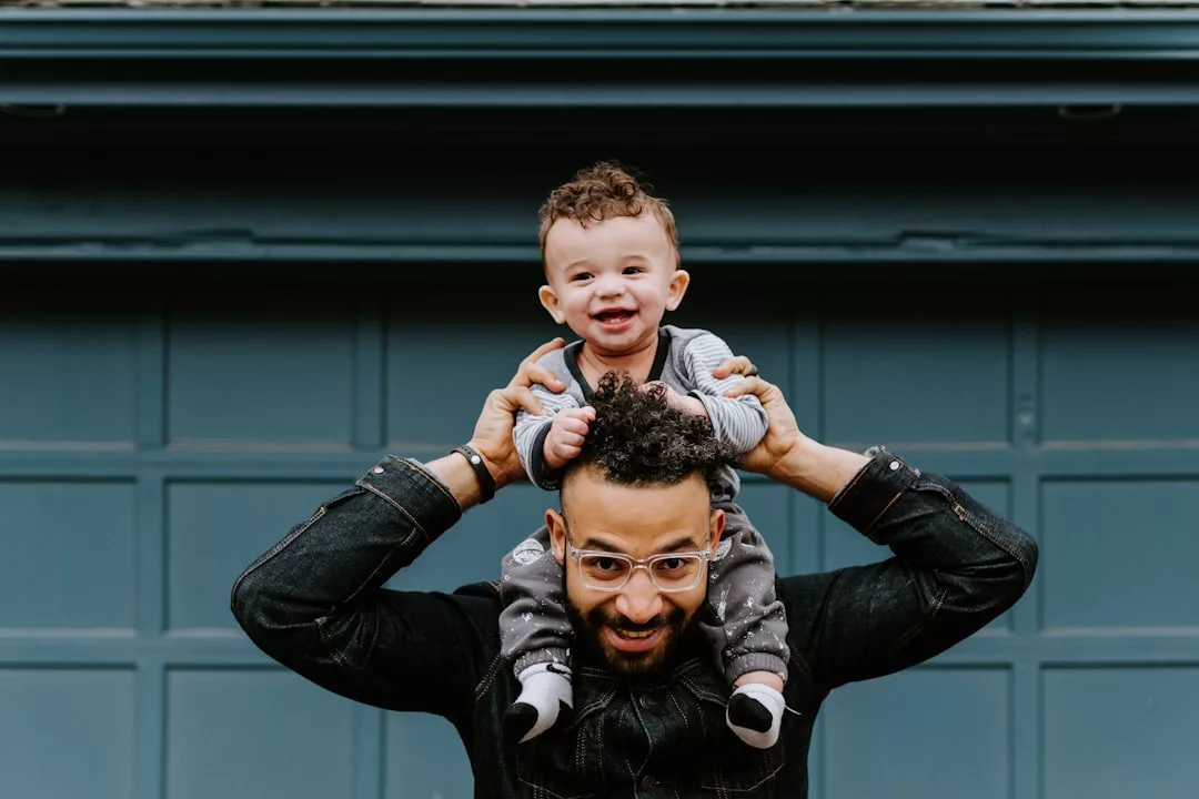 An adult wearing glasses holds a smiling baby on their shoulders in front of a blue garage door, capturing a joyful Glamesia lifestyle moment.