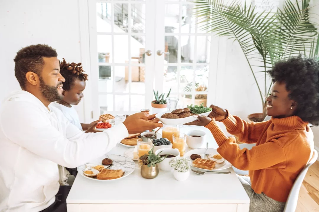 Three people sit at a white table, sharing food and smiling. The table is set with waffles, fruit, and drinks, creating a warm Laxary lifestyle vibe. Potted plants and sunlight fill the background with a touch of elegance.