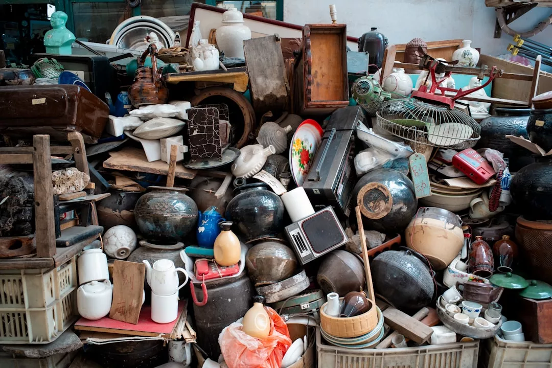 A large pile of assorted household items and antiques, including pottery, dishes, baskets, electronics, and kitchenware, stacked haphazardly in what appears to be a secondhand shop—where Glamesia meets forgotten luxury.