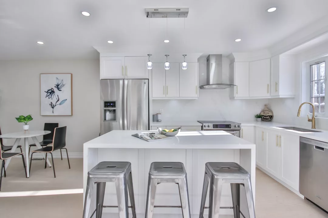 Modern white kitchen with stainless steel appliances, a center island with three metal stools, pendant lights, and a dining area—perfect for a stylish Glamesia lifestyle.