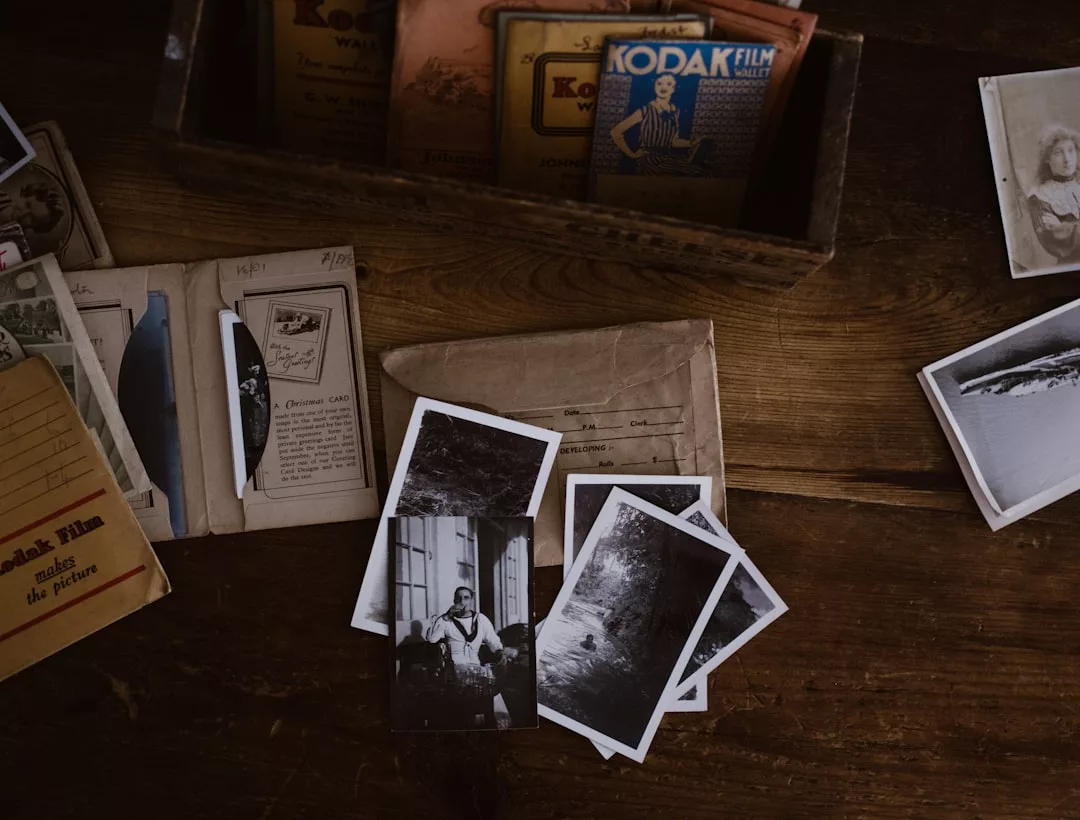Old black-and-white photographs, envelopes, and Kodak film boxes are spread out on a wooden table, capturing moments of lifestyle memories through images of people and landscapes.