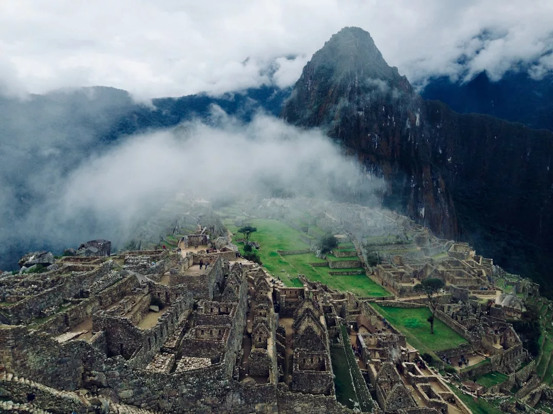 Ancient stone ruins of Machu Picchu sit atop a mountain, partially covered by mist, with a steep peak and cloudy sky in the background—an awe-inspiring scene that embodies the timeless allure and quiet luxury of Glamesia.