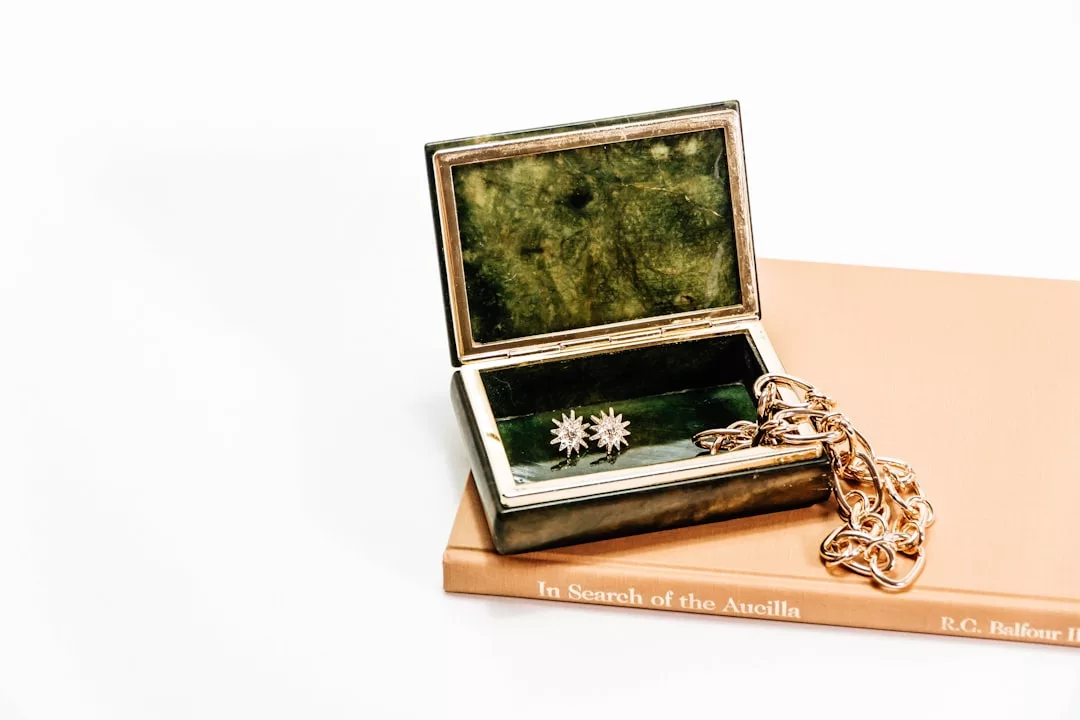 An open green Glamesia jewelry box with two flower-shaped earrings and a gold chain sits on a closed book titled "In Search of the Aucilla" by R.C. Balfour, radiating a touch of luxury.