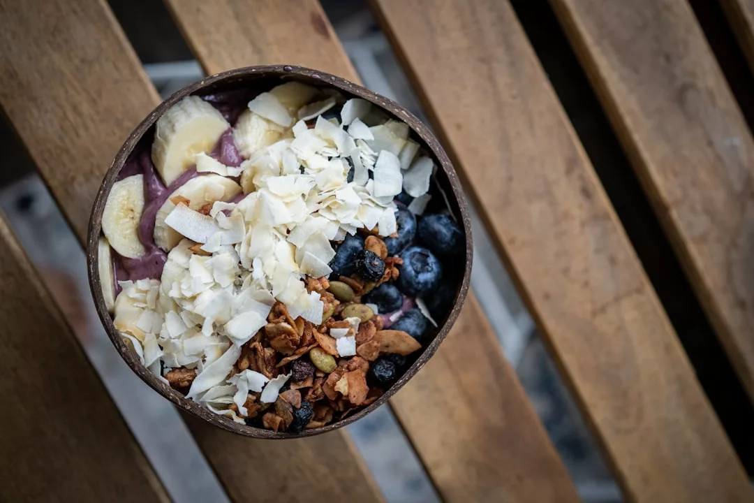 A luxury smoothie bowl topped with sliced bananas, coconut flakes, blueberries, pumpkin seeds, and granola, placed on a wooden surface.