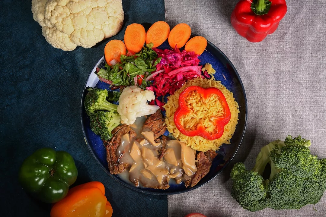 A plate with sliced carrots, leafy greens, shredded beets, broccoli, cauliflower, yellow rice with red bell pepper, and meat with brown gravy—perfect for a vibrant lifestyle inspired by Glamesia—surrounded by fresh raw vegetables.
