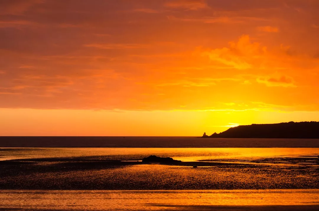 A bright orange sunset casts reflections over calm water and a silhouetted shoreline, with clouds partially covering the sky—a serene scene that captures the essence of luxury lifestyle in Glamesia.