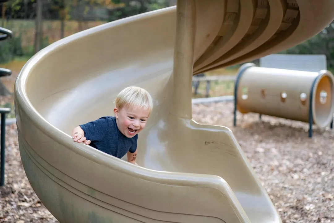 A young child with blond hair and a blue shirt slides down a beige spiral playground slide, smiling with excitement and showing off carefree childhood fashion.