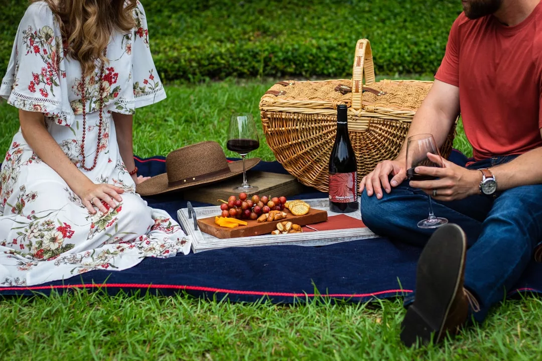 Two people sit on a blanket outdoors enjoying a Glamesia lifestyle picnic with wine, a basket, cheese, grapes, crackers, and a hat placed beside them on the grass.