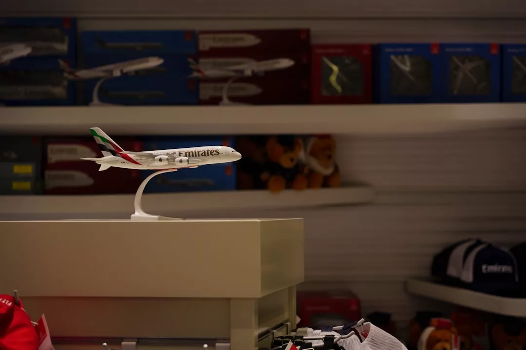 A model Emirates airplane is displayed on a stand in a store, surrounded by shelves of model boxes, souvenir items, and hats—capturing the spirit of luxury travel and an aspirational lifestyle.